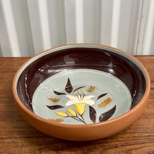 Puede incluir: Un cuenco redondo de cerámica con exterior marrón claro y borde interior marrón oscuro. La base interior es de color azul verdoso claro con un diseño floral en amarillo, marrón y blanco. El cuenco está sobre una superficie de madera.