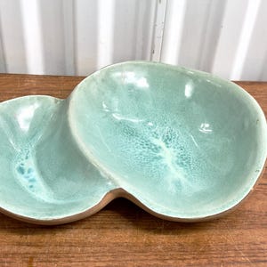 May include: A light green ceramic serving dish with two sections. The dish has a unique, organic shape and a speckled glaze.