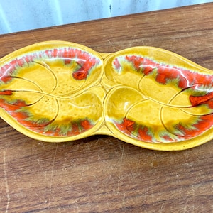 May include: A yellow ceramic serving dish shaped like two leaves with red and green accents. The dish has three sections.