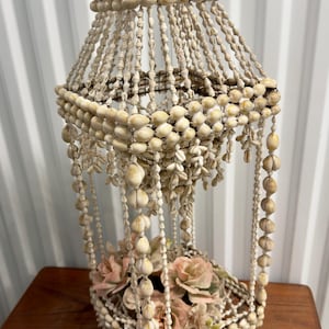 May include: A decorative hanging shell basket with a cone-shaped design. The basket is made of cream-coloured shells and features a square base filled with pink flowers. The shells are strung together to form the structure.