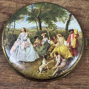 May include: A round, antique compact mirror with a painted scene of a gathering in a park. The scene features figures in period clothing, a dog, and trees. The compact has a gold-colored metal frame.