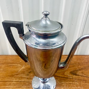 May include: A vintage silver coffee pot with a black handle and a shiny chrome finish. The pot has a round base and a tall, slender body.