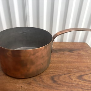 May include: A vintage copper pot with a single handle. The pot is round and has a hammered finish. It is sitting on a wooden surface.