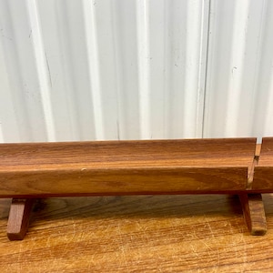 May include: A wooden bread slicer with a natural finish. The slicer has a rectangular base with a raised cutting surface and a vertical blade guide. The design includes two small legs for stability. The wood grain is visible.