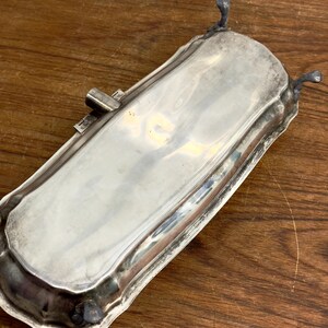 Puede incluir: Una bandeja de servir plateada con un borde decorativo y tres patas. La bandeja est&aacute; hecha de metal y tiene un acabado pulido.