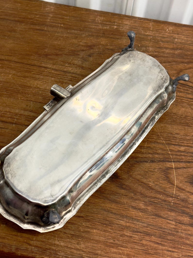 Puede incluir: Una bandeja de servir plateada con un borde decorativo y tres patas. La bandeja est&aacute; hecha de metal y tiene un acabado pulido.