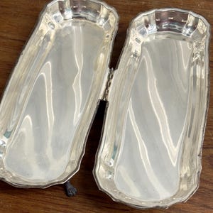 Puede incluir: Dos bandejas de servir plateadas con un borde decorativo y una conexi&oacute;n con bisagras. Las bandejas est&aacute;n abiertas y colocadas sobre una superficie de madera.