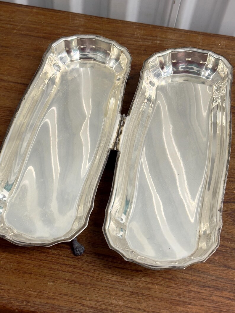 Puede incluir: Dos bandejas de servir plateadas con un borde decorativo y una conexi&oacute;n con bisagras. Las bandejas est&aacute;n abiertas y colocadas sobre una superficie de madera.