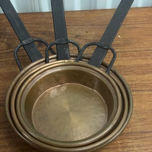 May include: A set of three copper pans with black metal handles. The pans are nested inside each other, showcasing their different sizes. The copper has a warm, reflective glow, and the handles have a loop for hanging.