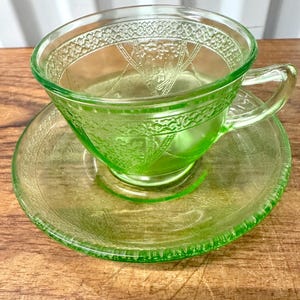 Può includere: Un set composto da una tazza da tè e un piattino abbinato in vetro verde chiaro traslucido. La tazza presenta un delicato motivo in rilievo di uccelli e fogliame all'esterno, con un bordo testurizzato. Il piattino ha un motivo decorativo coordinato.