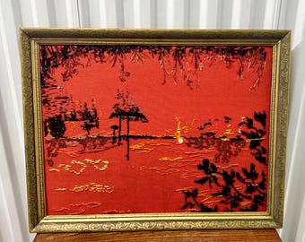 May include: A framed artwork featuring a sunset scene. The artwork has a red background with black and yellow embroidered details depicting trees, water reflections, and a golden frame. The frame has an ornate design.