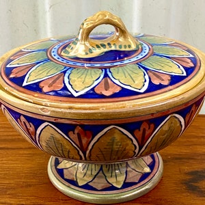 Może przedstawiać: Ceramiczna miska z pokrywką, ozdobiona ręcznie malowanym wzorem stylizowanych liści w odcieniach zieleni, brązu, błękitu i różu. Miska ma podstawę w kształcie cokołu i złoty uchwyt na pokrywce.