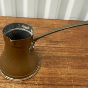 May include: A copper-colored Turkish coffee pot with a long, olive-green handle. The pot has a wide base that tapers to a narrow opening. The handle is attached to the side of the pot and has a small hole at the end.