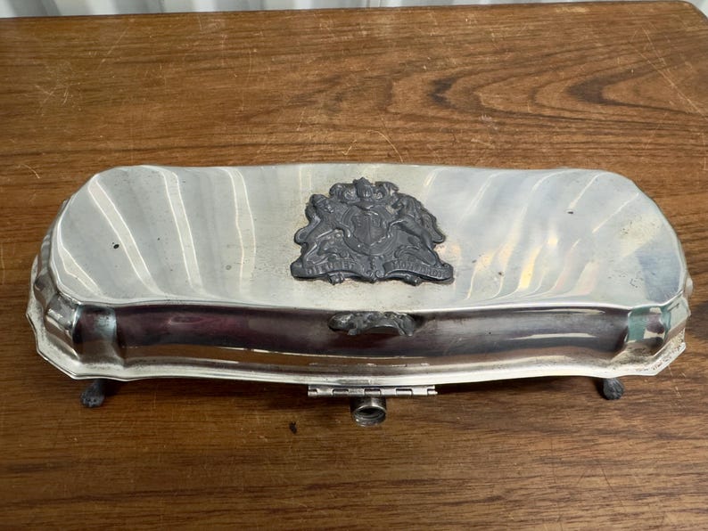 Puede incluir: Una caja de alhajas plateada con una tapa con bisagras y patas decorativas. La caja presenta un escudo negro y blanco con una corona en la parte superior.