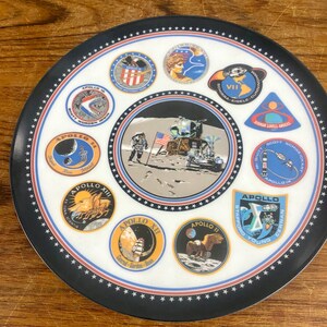 May include: A white ceramic plate with a black border and a red and blue trim. The plate features a central image of the Apollo 11 lunar module landing on the moon, surrounded by nine circular patches representing different Apollo missions. Each patch features the mission number, crew names, and a unique design.