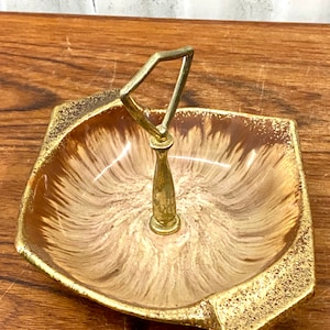 Op de afbeelding: Een bruine en gouden keramische schaal met een gouden metalen handvat. De schaal heeft een swirl-patroon en een verhoogde rand.