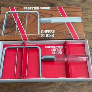 Op de afbeelding: Een zilveren kaassnijder met een transparante acryl handvat. De snijder zit in de originele verpakking, die wit is met rode accenten en de woorden "Frontier Forge Cheese Slicer" erop gedrukt.