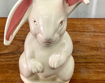 May include: A white ceramic bunny figurine with pink ears. The bunny is sitting with its paws tucked in front of it.