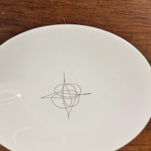 May include: A white, round plate with a minimalist design. A gray, abstract line drawing is centered on the plate. The plate is set against a wooden surface.