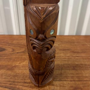 Könnte beinhalten: Eine geschnitzte Tiki-Statue aus Holz mit detailliertem Gesicht und Körper. Die Statue ist braun mit eingelegten irisierenden Augen. Der Tiki steht auf einer Holzoberfläche.