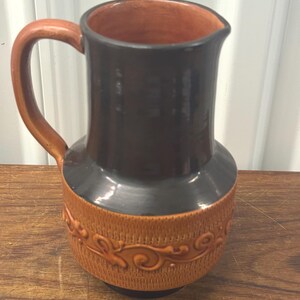 May include: A brown and black ceramic pitcher with a handle. The top and handle are brown, while the body is black and brown with a decorative pattern. The pitcher is likely used for serving beverages.