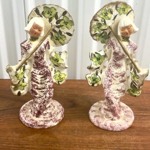 May include: Two ceramic figurines of women in traditional attire, each holding a parasol. The figures have white hair and are dressed in cream and burgundy robes. The parasols are cream-colored with green floral designs.