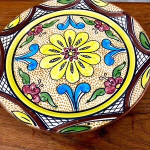 May include: A large, round ceramic plate with a brown, yellow, blue, and red floral design. The plate has a raised rim and a scalloped edge.
