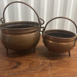 May include: Two copper-colored cauldrons with arched handles and three small legs. The cauldrons are different sizes, with the larger one on the left and the smaller one on the right. The cauldrons are sitting on a wooden surface.