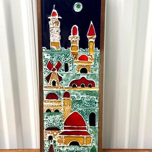 May include: A rectangular ceramic tile with a dark blue sky background and a cityscape design. The buildings have red, yellow, and white accents, with a green textured base. The tile is framed in a dark wood.