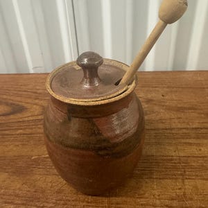 Puede incluir: Un tarro de miel de cerámica de forma redonda, con tonos marrones y rojizos, que incluye una tapa a juego con un pomo central. Un cucharón de miel de madera clara está insertado en una pequeña muesca de la tapa, sobresaliendo.
