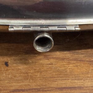 Puede incluir: Una petaca de metal plateado con una tapa con bisagras y un pico redondo. La petaca est&aacute; sobre una superficie de madera.