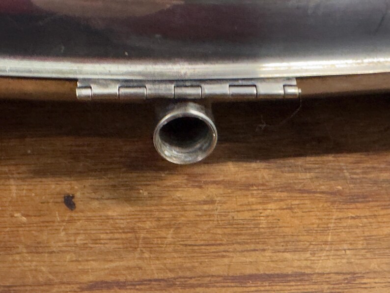 Puede incluir: Una petaca de metal plateado con una tapa con bisagras y un pico redondo. La petaca est&aacute; sobre una superficie de madera.