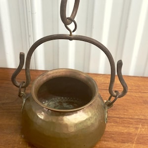 May include: A hammered brass cauldron with a curved handle and hanging ring. The metal pot has a rounded body and a dark interior. The handle and hanging hardware are made of a dark, aged metal. The cauldron is suspended above a wooden surface.