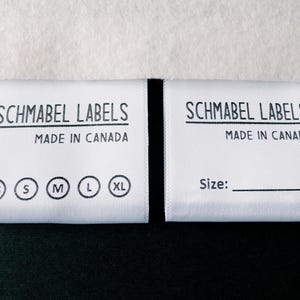 May include: Two white fabric labels with black text. The label on the left says "Schmabel Labels Made in Canada" and has size options: XS, S, M, L, XL. The label on the right says "Schmabel Labels Made in Canada" and has the word "Size:" printed on it.