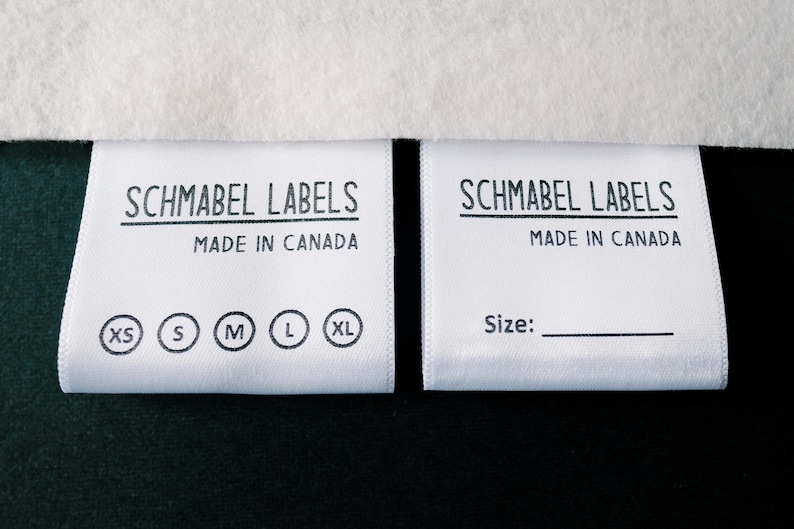 May include: Two white fabric labels with black text. The label on the left says "Schmabel Labels Made in Canada" and has size options: XS, S, M, L, XL. The label on the right says "Schmabel Labels Made in Canada" and has the word "Size:" printed on it.