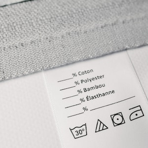 May include: A white fabric care label with black text and symbols. The label lists the fabric composition: % Cotton, % Polyester, % Bamboo, % &Eacute;lasthanne. It also includes washing instructions: 30 degrees Celsius, a triangle with lines inside, a circle with a dot inside, and an iron with a dot inside.
