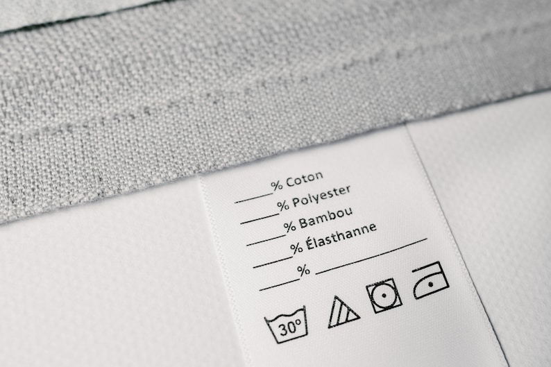 May include: A white fabric care label with black text and symbols. The label lists the fabric composition: % Cotton, % Polyester, % Bamboo, % &Eacute;lasthanne. It also includes washing instructions: 30 degrees Celsius, a triangle with lines inside, a circle with a dot inside, and an iron with a dot inside.
