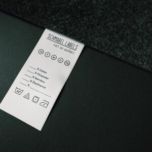 May include: A white clothing label with black text and symbols. The label reads "Schmabel Labels Fait au Quebec" and includes a size guide with the letters "TP, P, M, G, TG". The label also includes a list of fabric percentages: "Coton, Polyester, Bambou, Elasthanne". The label includes washing instructions with a 30 degree symbol, a triangle, a circle with a dot in the centre, and a square with a circle inside.