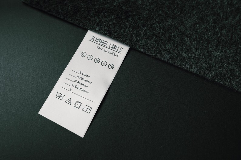 May include: A white clothing label with black text and symbols. The label reads "Schmabel Labels Fait au Quebec" and includes a size guide with the letters "TP, P, M, G, TG". The label also includes a list of fabric percentages: "Coton, Polyester, Bambou, Elasthanne". The label includes washing instructions with a 30 degree symbol, a triangle, a circle with a dot in the centre, and a square with a circle inside.
