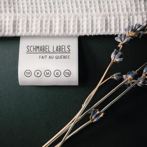 May include: A white fabric label with black text that reads "Schmabel Labels Fait au Qu&eacute;bec" and size indicators: TP, P, M, G, TG. The label is on a dark green surface with dried lavender sprigs.