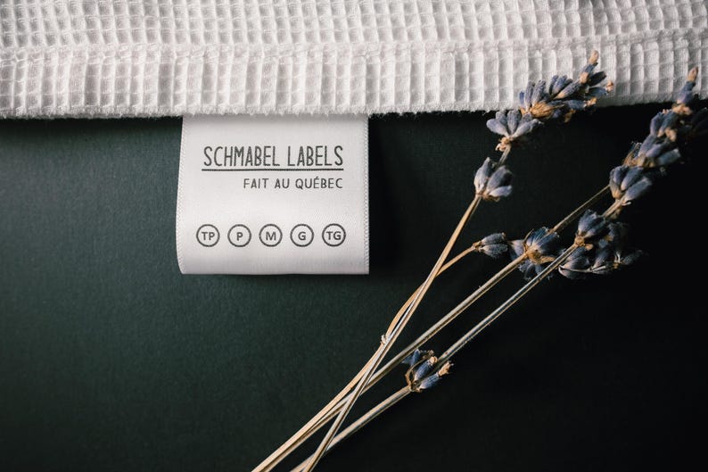 May include: A white fabric label with black text that reads "Schmabel Labels Fait au Qu&eacute;bec" and size indicators: TP, P, M, G, TG. The label is on a dark green surface with dried lavender sprigs.