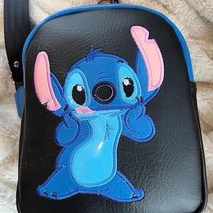 May include: A black faux leather crossbody bag with a blue and pink embroidered Stitch character from the Disney movie Lilo & Stitch.