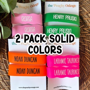May include: Four sets of two silicone wristbands in pink, green, orange, and hot pink, each featuring custom names printed in white or black. The bands are displayed on white cards labeled with the brand name the Peachy Orange.
