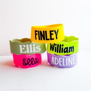May include: Four colorful pet bowls stacked on top of each other. The bowls are yellow, green, pink, and purple. Each bowl has a different name printed on it in a contrasting color: Finley, Ellis, William, and Adeline.