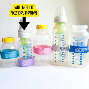 May include: Five clear plastic baby bottles with colourful silicone bands featuring names. The second bottle from the left has a white cap and a black arrow pointing to it with the yellow text WILL NOT FIT 4OZ DR. BROWN. The bands are blue, purple, pink, grey, and blue.