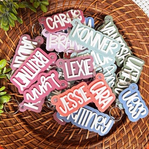 May include: A collection of colorful personalized keychains with names written in white lettering. The keychains are made of a soft, flexible material and have a glossy finish.