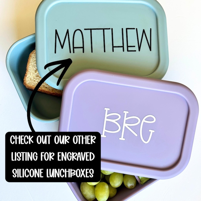 May include: Two rectangular silicone lunchboxes with lids. The top light blue lid features the black text MATTHEW. The bottom lavender lid features the white text BRG. A black arrow points from a black text box to the top lid.