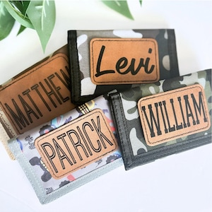 May include: Four personalized wallets with names: "Matthew", "Levi", "Patrick", and "William". The wallets feature a camouflage pattern and a leather patch with the name in black lettering. The wallets are in shades of green, gray, and tan.