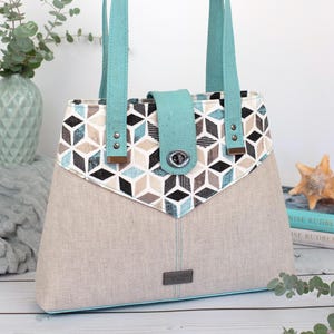May include: A structured tote bag featuring a beige fabric base and a top panel with a geometric cube pattern in shades of black, white, beige, and teal. It includes teal straps, a matching teal flap with a silver twist-lock, and a small metal tag.