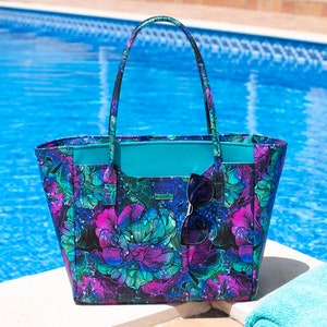 May include: A large tote bag with a floral print in shades of purple, blue, and green. The bag has a turquoise lining and a pair of sunglasses are tucked into the top of the bag.